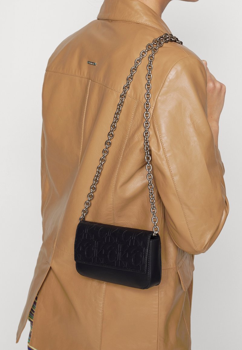 Borsa a spalla in pelle nera con motivo in rilievo del logo e tracolla in catena argentata indossata sopra un giubbotto di pelle beige.