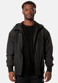 Schwarze, leichte Jacke mit Kapuze, Reißverschluss, elastischen Bündchen und strukturiertem Stoff. Mit Logo auf der linken Brust und seitlichen Taschen.