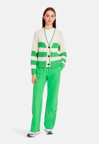 Cremefarbener Cardigan mit grünen Streifen, strukturiertem Strick und fünf Knöpfen. Kombiniert mit leuchtend grünen Hosen und gemusterten Turnschuhen.