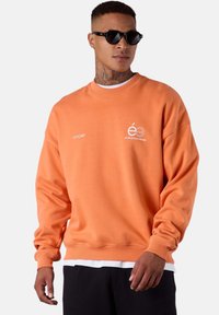 Sweatshirt orange avec poignets et ourlet côtelés, arborant des logos blancs à l'avant. Texture lisse, avec un col rond, superposé à une chemise blanche.