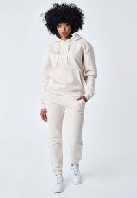 Felpa crema con tasca frontale e lacci, abbinata a pantaloni sportivi coordinati con tasche laterali e orli a coste, indossata con sneakers bianche.