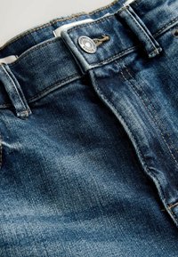 Mörkblå denimjeans med en platt midja, silverknapp som stängning och kontrasterande guldtråd synlig på sömmarna.