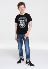 Black cotton t-shirt featuring a Hogwarts crest design in white; paired with blue denim jeans and black sneakers with white stripes.
