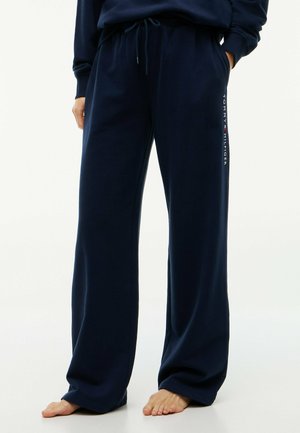 Personne portant un pantalon de jogging bleu marine à jambes larges avec une taille à cordon et le logo Tommy Hilfiger sur la jambe, pieds nus, mains dans les poches.