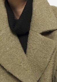 Beige uldblandingsfrakke med en tekstureret overflade, der har en dyb sort ribstrikket krave. Frakken har et struktureret, skræddersyet design.