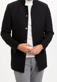 Cappotto di lana nera con colletto alto, design a abbottonatura semplice, sei bottoni e una texture liscia. Indossato sopra un maglione bianco.