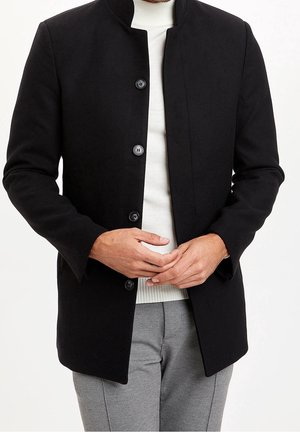 Svart ullkappa med hög krage, enfärgad design, sex knappar och en mjuk textur. Bärs över en vit tröja.
