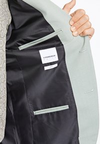 Rokās turēts gaismas pelēks žakete ar atvērtu priekšpusi, kas rāda melnu acs auduma oderi un divas iekšējās kabatas ar Lindbergh White slim fit etiķeti.