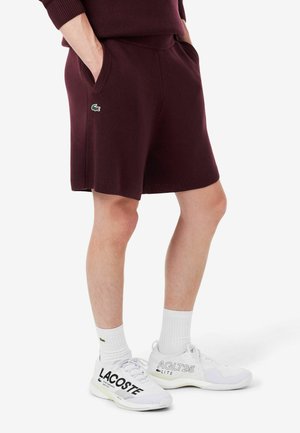 Unterkörper, der dunkelrote Shorts trägt, die Hände in den Taschen, weiße Socken und weiße Lacoste-Sneaker mit schwarzer und grauer Schrift.