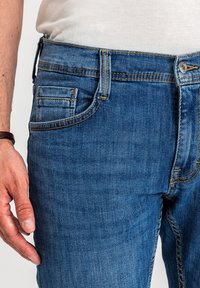 Zbliżenie osoby noszącej niebieskie dżinsy z żółtym przeszyciem i białą koszulę, pokazujące lewą rękę i opaskę na nadgarstek.