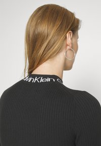 Zwarte geribde trui met een ronde halslijn en een contrasterende witte logoband, die textuur en aansluitend ontwerp benadrukt.