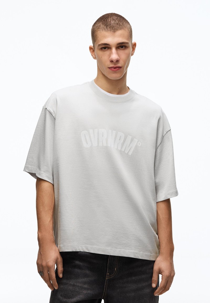 Jeune homme aux cheveux courts portant un T-shirt gris clair ample avec le texte "OVRNRM" et un jean foncé, debout devant un fond blanc.
