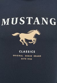 Tmavě modré tričko s velkým bílým nápisem "MUSTANG", zlatým siluetou koně a dalšími brandingovými detaily v bílé a zlaté barvě.