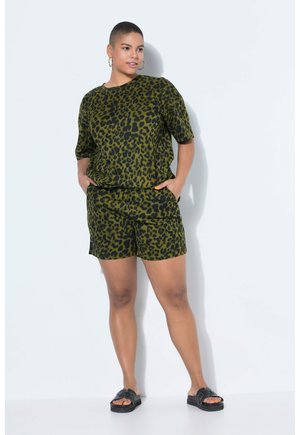 Femme debout, les mains dans les poches, portant un haut à manches courtes imprimé léopard vert et noir et un short assorti, avec des sandales noires à enfiler.