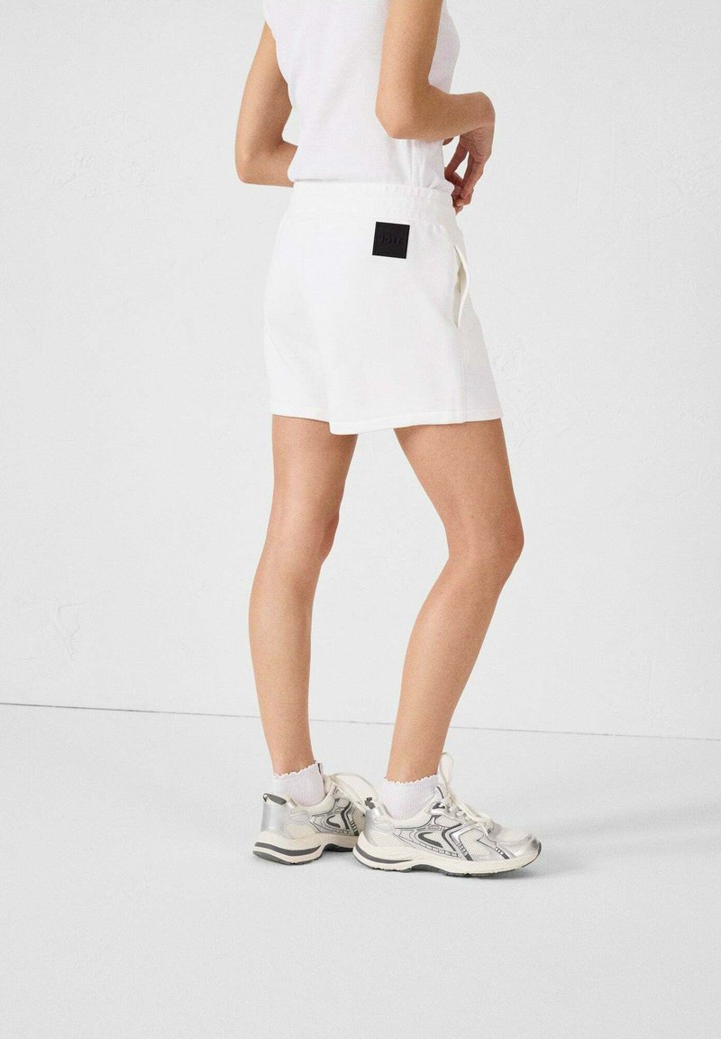Pantaloni corti bianchi in cotone con vita elasticizzata, dotati di una tasca posteriore e una patch del logo nera. Abbinati a scarpe da ginnastica argento e bianche.