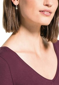 Silvriga hängande örhängen med en päronformad sten, bärs med en djupt bordeauxfärgad V-ringad topp som framhäver en slät halslinje och kort hår.