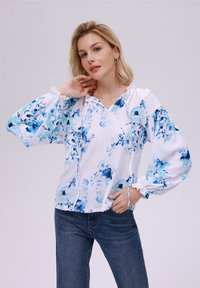 Blusa bianca con motivo floreale blu, maniche a sbuffo con polsini elasticizzati, scollatura con cordoncino, tessuto testurizzato. Abbinata a jeans in denim scuro.