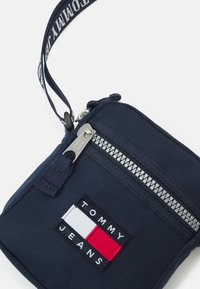Pochette a tracolla in tessuto blu navy con zip argentata, caratterizzata da un logo brandizzato in rosso, bianco e blu. Include una tracolla staccabile.