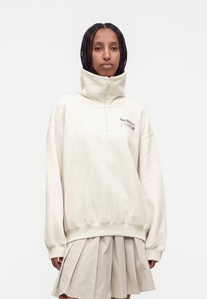 Mujer con el pelo trenzado que lleva una sudadera blanca de New Balance con cuello alto y una falda plisada beige sobre un fondo liso.