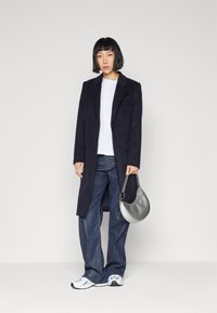 Marineblå uldfrakke med slåede klapper, hvid t-shirt, brede blå denim jeans og grå håndtaske; modellen bærer hvide og grå sneakers.