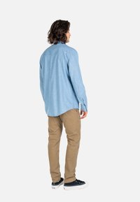 Hombre con cabello rizado que lleva una camisa de manga larga azul claro, pantalones beige y zapatillas negras, de pie de espaldas sobre un fondo blanco.