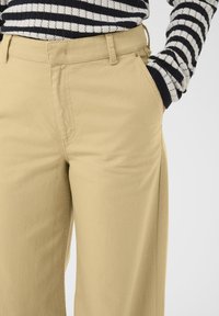 Pantalon en coton beige à coupe décontractée, avec des poches avant et des coutures visibles. L'arrière-plan est blanc uni.