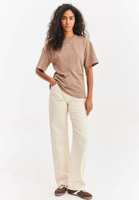 Kobieta z długimi, kręconymi włosami, ubrana w brązową, oversize'ową koszulkę, kremowe spodnie z szerokimi nogawkami oraz brązowe trampki, stojąca z jedną ręką w kieszeni.