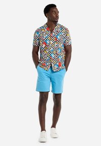 Kurzarmhemd mit einem bunten Cartoon-Muster auf einem schwarz-weißen Schachbrett-Hintergrund, kombiniert mit hellblauen Shorts und weißen Sneakern.