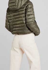 Olivengrüne Steppjacke mit horizontalem Steppdesign und Kapuze, kombiniert mit hellbeigen, geraden Hosen. Glatte, glänzende Textur.