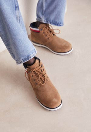 Lysebrune ruskindsstøvler med hvide såler og rødt, hvidt og blåt bånd, båret med lyseblå denimbukser på en beige gulv.