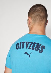Zils T-krekls ar īsām piedurknēm, uz kura aizmugures ir uzraksts "CITYZENS" tumši z blue krāsā. Dizainā ir iekļauta Puma logo zem uzraksta.