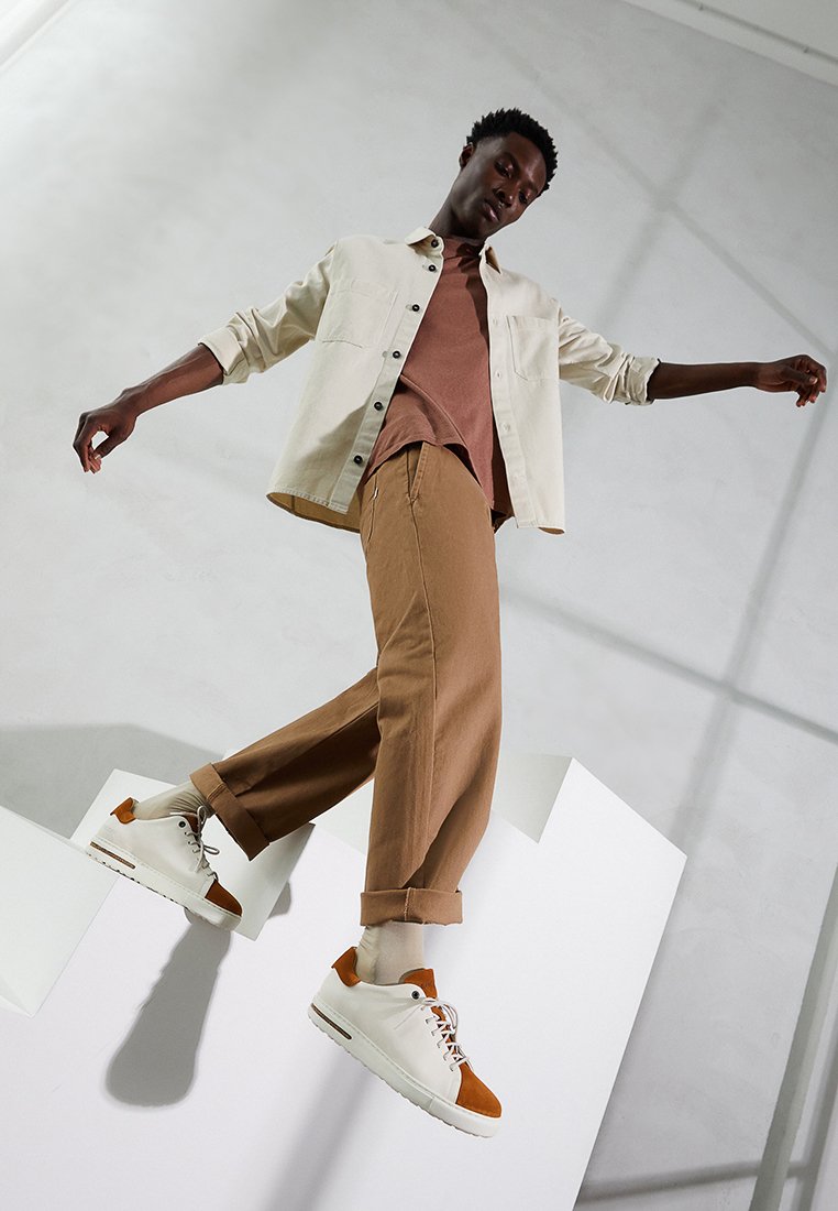 Lässiges Outfit bestehend aus einem cremefarbenen Hemd mit Knöpfen, einem rostfarbenen T-Shirt, braunen weiten Hosen und weißen Sneakers mit orangen Akzenten.