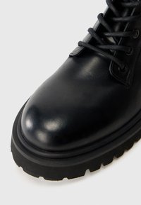 Stivale in pelle nera con punta rotonda, superficie testurizzata e suola in gomma spessa con bordo seghettato. I lacci si estendono attraverso gli occhielli.