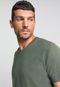 Donkergroene katoenen t-shirt met een ronde hals, korte mouwen en een klein zilveren logo op de borst. Glad van textuur.