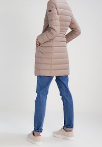 Vadderad beige jacka med horisontell quiltad design, har en hög krage och en logotyp på ärmen, parat med blå jeans och ljusa sneakers.