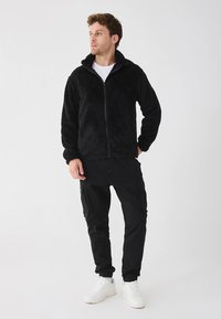 Musta fleece-vetoketjullinen takki, jossa korkea kaulus, yhdistetty mustiin cargo-housuihin ja valkoisiin tennareihin. Sileä pinta, rento muotoilu, väljä istuvuus.