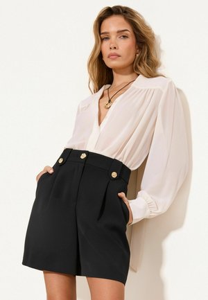 Femeie purtând o bluză albă cu mâneci lungi băgată în pantaloni scurți negri cu talie înaltă și nasturi aurii, stând în fața unui perete alb.