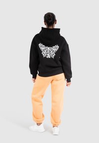 Felpa con cappuccio nera con un design di farfalla floreale bianca sulla schiena, abbinata a pantaloni della tuta arancioni larghi e sneakers bianche.