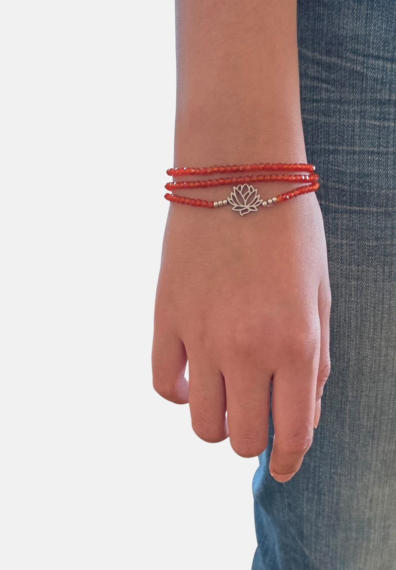 Poignet portant un bracelet triple tour en perles rouges avec un pendentif lotus en argent, à côté d'un pantalon en denim bleu.