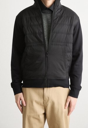 Man draagt een zwart jack met ritssluiting met gestructureerde voorkanten over een donkere trui en beige broek.