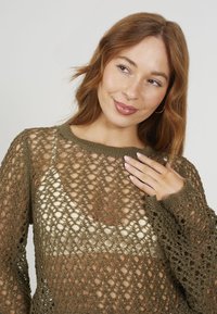 Oliwkowy sweter dzianinowy o luźnym, otwartym splocie, szerokich rękawach i ściągaczu przy dekolcie, nałożony na jasną bluzkę.