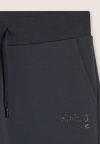 Un pantalon de survêtement gris foncé avec une taille côtelée, un cordon de serrage ajustable et un logo décoratif brillant en strass sur la cuisse.