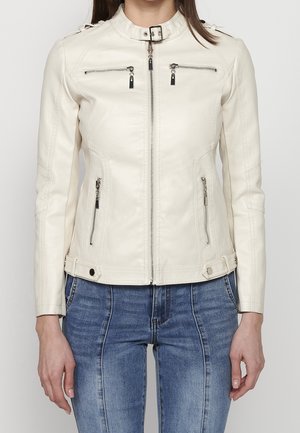 Femme portant une veste en cuir blanc cassé avec poches zippées et col à boucle, associée à un jean bleu, visible du cou aux hanches.