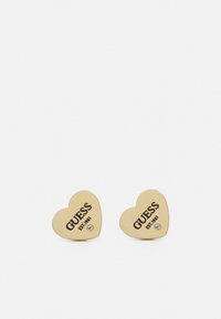 Gouden hartvormige oorbellen met zwarte "GUESS EST. 1981" tekst, voorzien van een kleine heldere steentjesaccent op elke oorbel.