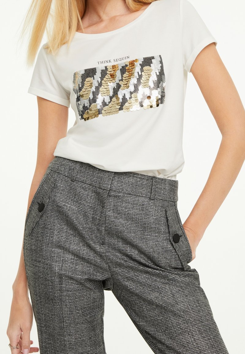 Femme portant un t-shirt blanc avec un motif de sequins dorés et argentés et un pantalon taille haute à carreaux gris avec des accents de boutons.