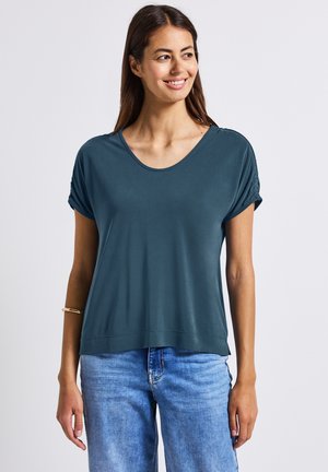 Haut court en teal foncé à col rond et coupe décontractée. Texture lisse et plis subtils au niveau des manches, associé à un jean bleu clair.