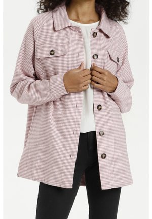 Femme portant une veste boutonnée à motif pied-de-poule rose et blanc par-dessus un haut blanc et un pantalon noir, ajustant la veste avec les deux mains.