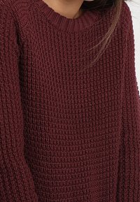 Bordowy, dziergany sweter z fakturowanym wzorem, okrągłym dekoltem i luźnym krojem. Materiał wydaje się miękki i ciepły, z wyraźnym prążkowaniem.