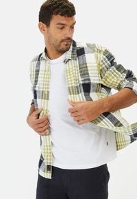 Koszula flanelowa w kratę, w zielone, granatowe i białe wzory; styl zapinany na guziki; postrzępione krawędzie; noszona na białej koszulce, stylizacja casualowa.