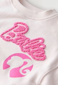 Lyserød sweatshirt med tekstureret lyserød brodering, der stavr "Barbie" og et cirkulært logo med en profil-silhuet i en matchende nuance.